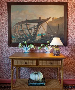 Hallway with red and white, floral traditional wallpaper pattern, wooden console table with wicker draws and low shelf, decorated with table lamp, flowers and books, large painting with dark wood frame hung above console table, red painted skirting board and patterned carpet