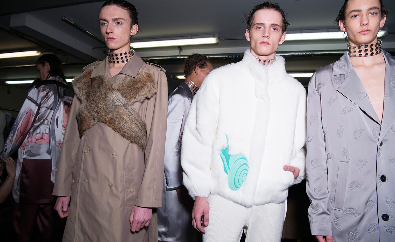 Five male models wearing looks from JW Anderson&#039;s collection. One model is wearing a beige coat with X-shaped fur panel across the chest. Another model is wearing white pants and a white short fur jacket with blue detail. And the other three models are wearing grey pieces, one with a pink and brown design and another with a repeating pattern. All models are wearing thick clear chokers with metal studs