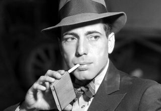 Posed portrait of actor Humphrey Bogart (1899-1957) wearing a hat and bow tie with a pack of cigarettes, for Warner Bros Studios, 1938.