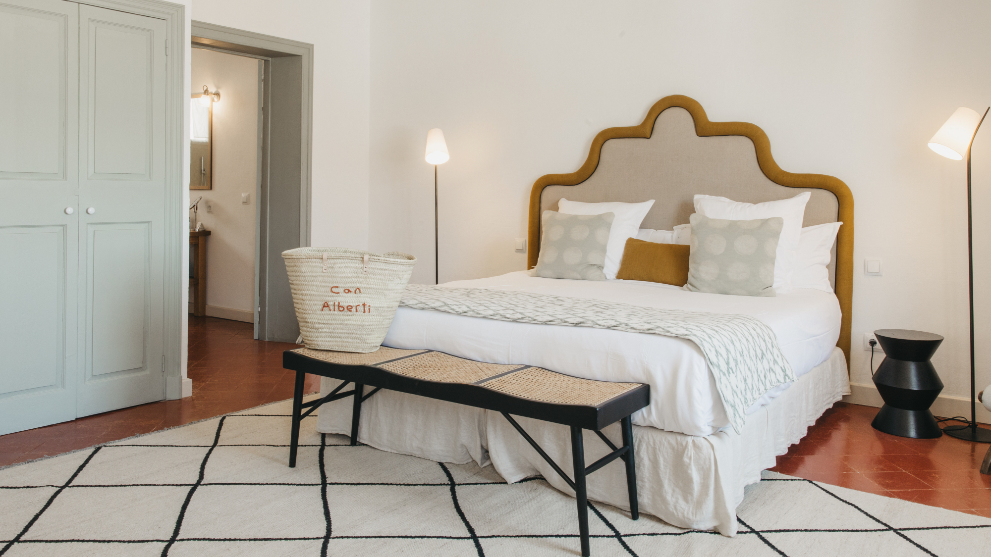 Double bed with padded headboard stands on a grid-patterned rug on a terracotta tiled floor, with a grey-green built-in wardrobe, in the Can Alberti 1740 hotel in Mahon, Menorca