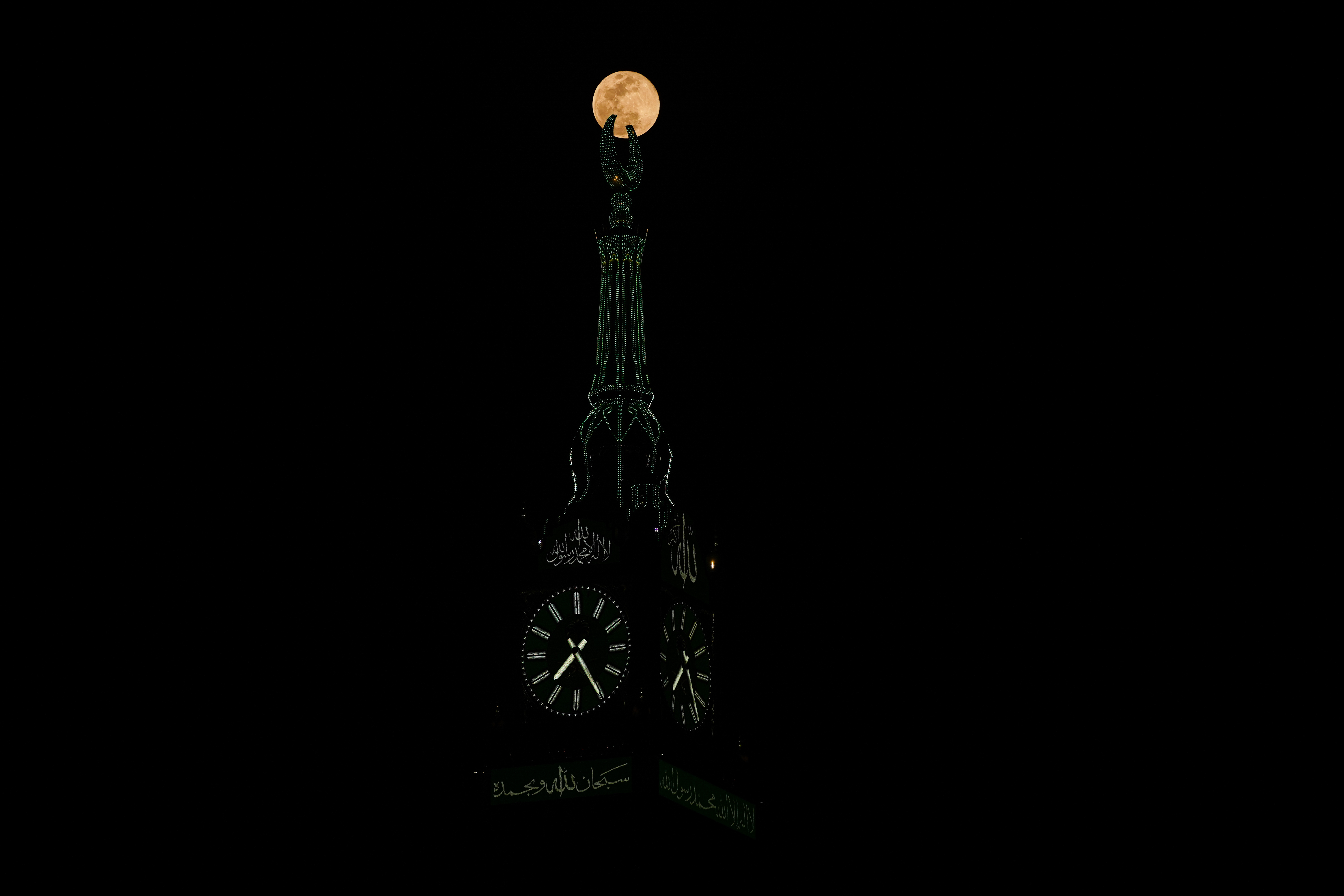 The full lunar disk shines at the apex of an ornate, illuminated clock tower in a dark night sky.
