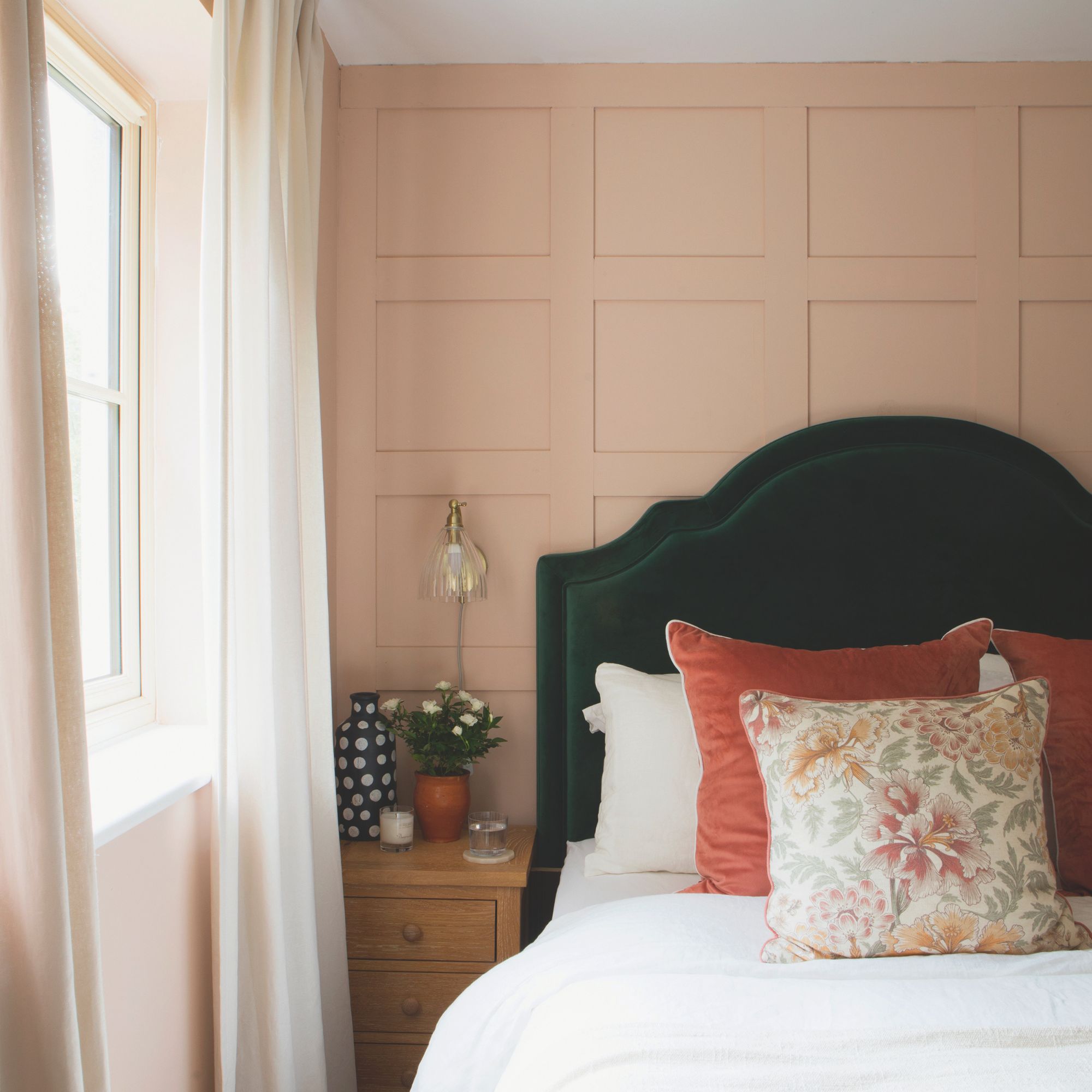 Bedroom with pink wall panelling behind a bed, and a window with sheer curtains to the left