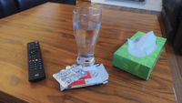 A tall glass of water, an open box of ibuprofen with a partially used blister pack, a remote control, and a box of tissues sit on a wooden coffee table.