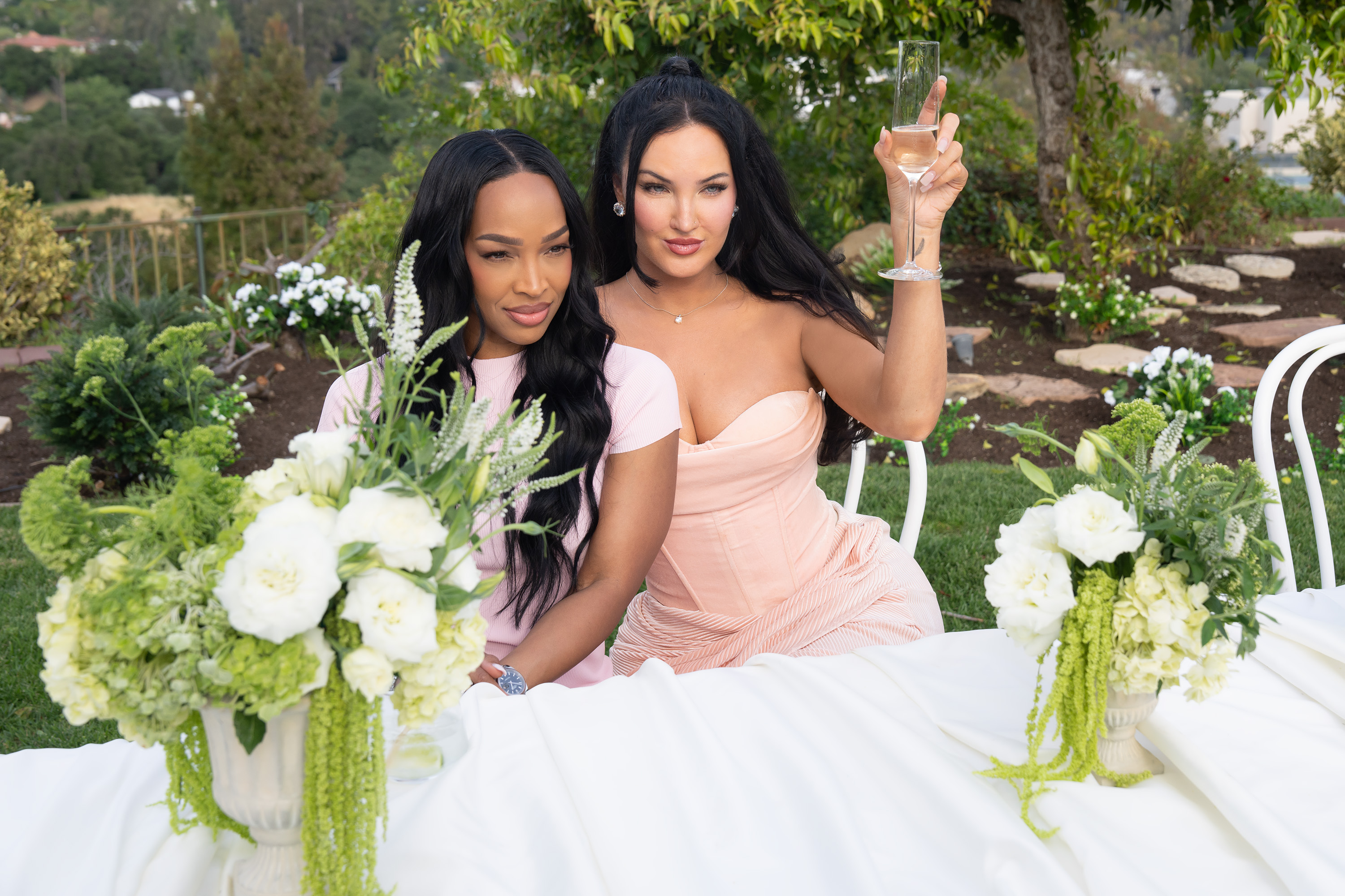 MALIKA HAQQ and NATALIE HALCRO sit at a white table cloth table outdoors while holding glasses of wine in a still from the reality show the girls