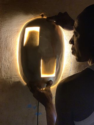 An African man installing an oval shaped handcrafted light onto a wall.