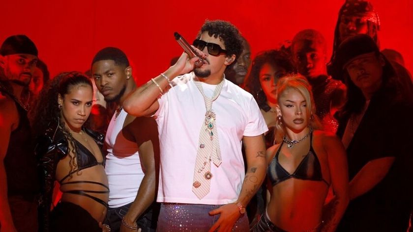 Bad Bunny on stage in a white t-shirt against a red backdrop surrounded by backing dancers.