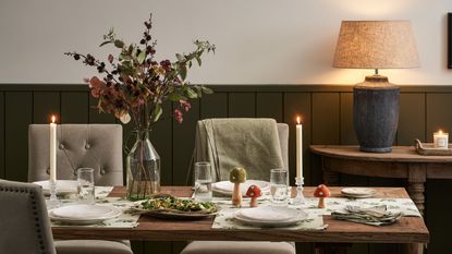 A wooden dinner table with an autumnal tablescape, a vase of dried stems, and a table lamp on in the background
