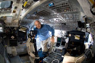 a bald man in a blue flight suit floats inside a spacecraft