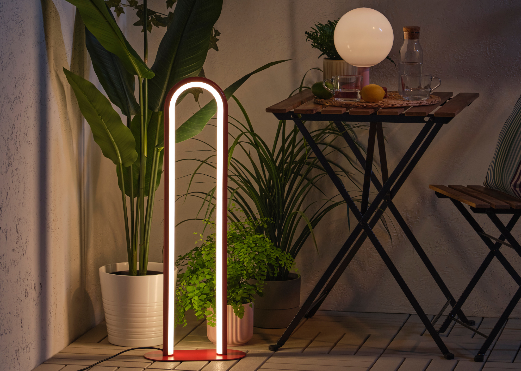 IKEA's SOLVINDEN outdoor floor lamp in a red arc design next to a small table and chair in a garden corner at dusk with plants and foliage against the white walls