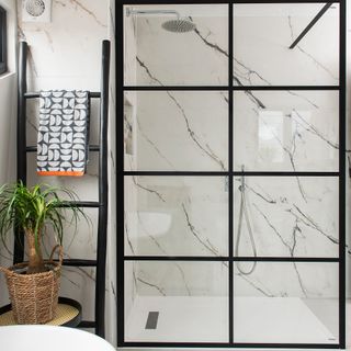 A modern monochrome bathroom with black Crittal-style shower enclosure, large marble-effect tiles, indoor houseplant, black ladder and black, orange and grey towel with geometric pattern