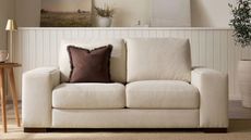 beige sofa with chocolate brown throw pillow, timber side table with gold lamp, and panelling on the wall behind with artwork