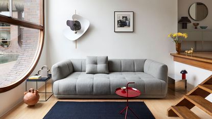 Sofa trends 2023. HAY Quilton sofa, gray fabric, sunken living room with large window, wooden flooring, artwork on wall, blue rug, red side table