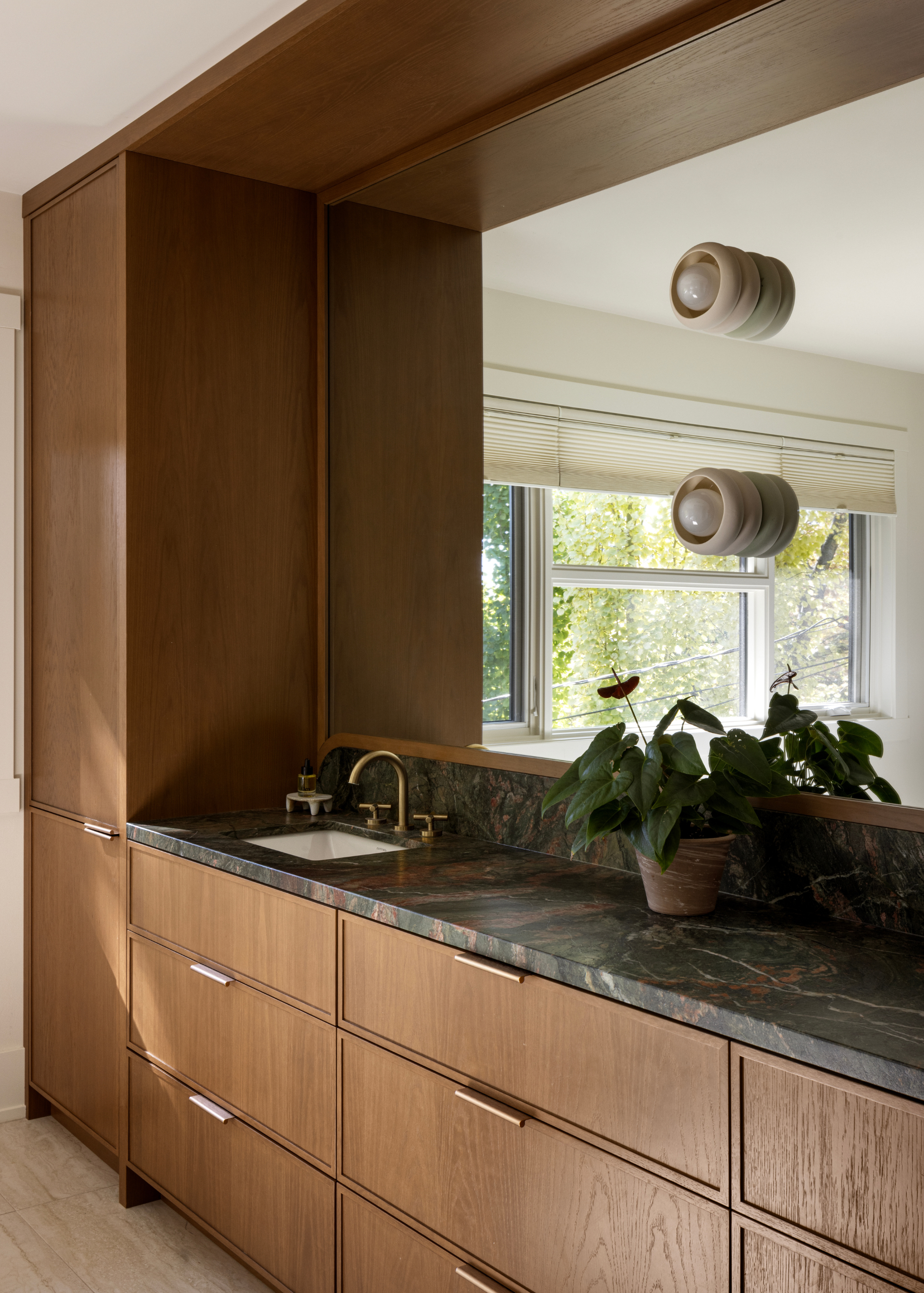 A bathroom with a dark marble counter, his and hers sinks, a potted plant, wooden cabinets and drawers, light sconces on the mirror, and a large window