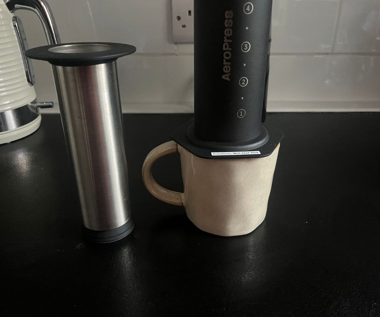 A black AeroPress on top of a beige coffee mug, with the stainless steel plunger beside it.