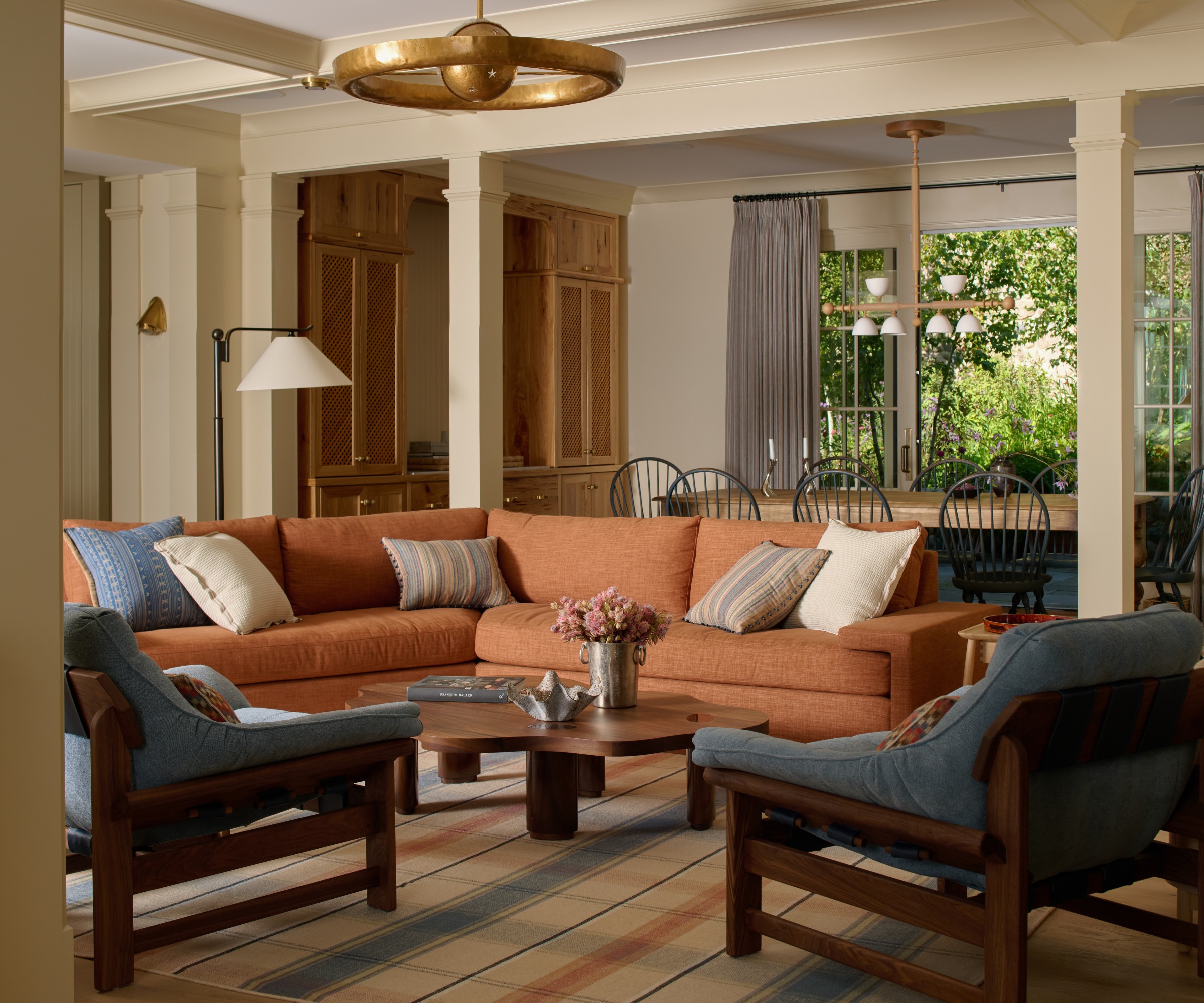 a living room with a large burnt orange couch, two blue accent chairs, a matching rug, and beams that separate the living area from the dining room behind