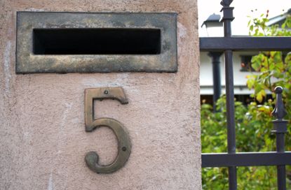 A mail slot set into a house pillar with a wrought iron fence and a brass number five.