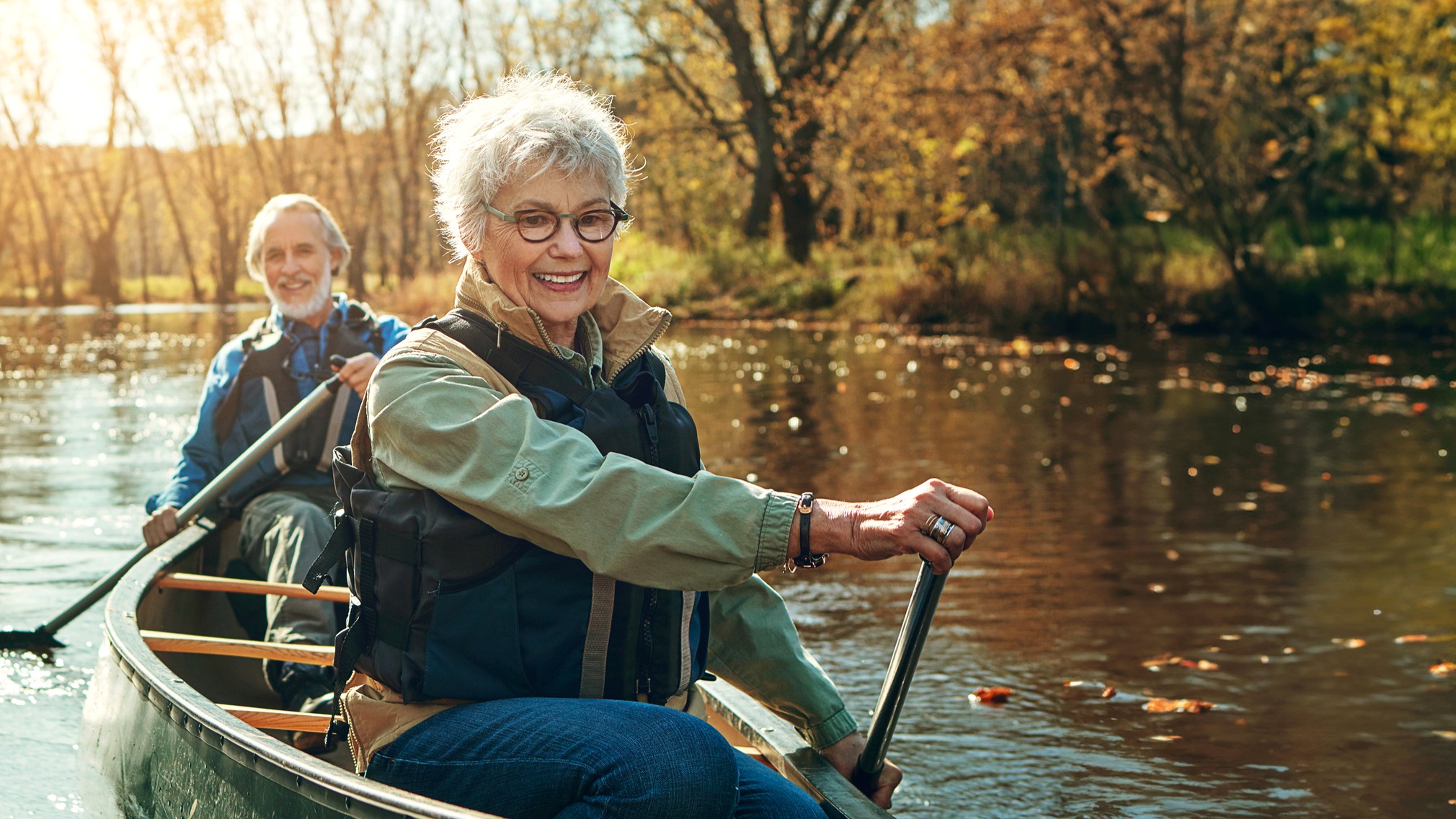 A mature couple in their 70s paddles a canoe on a fall day. They look happy and calm.