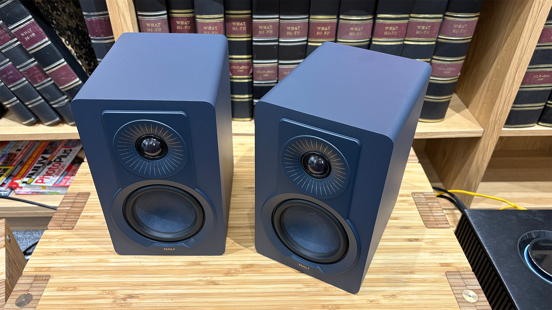 Dali Kupid standmount speakers on wooden AV rack in front of bookcase, slight top-down angle