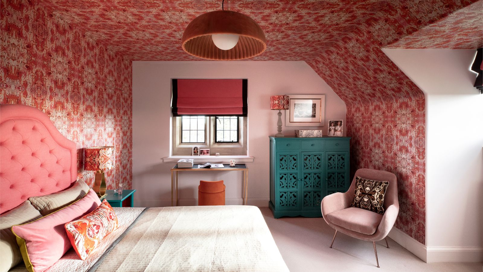 A pink wallpapered attic bedroom, with a white accent wall. A pink upholstered bed with cream bedding on it, and a blue painted dresser in the corner. 