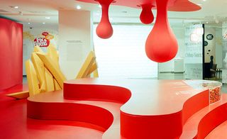 Currywurst museum, white floor and ceiling with spotlights, red curved platform and red drips suspended from the low ceiling, with yellow padded strips, to simulate giant chips and curry sauce