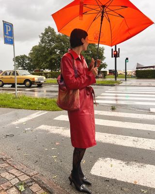 A woman wears a red leather trench coat, tights, black slingbacks, and a sequin shoulder bag.