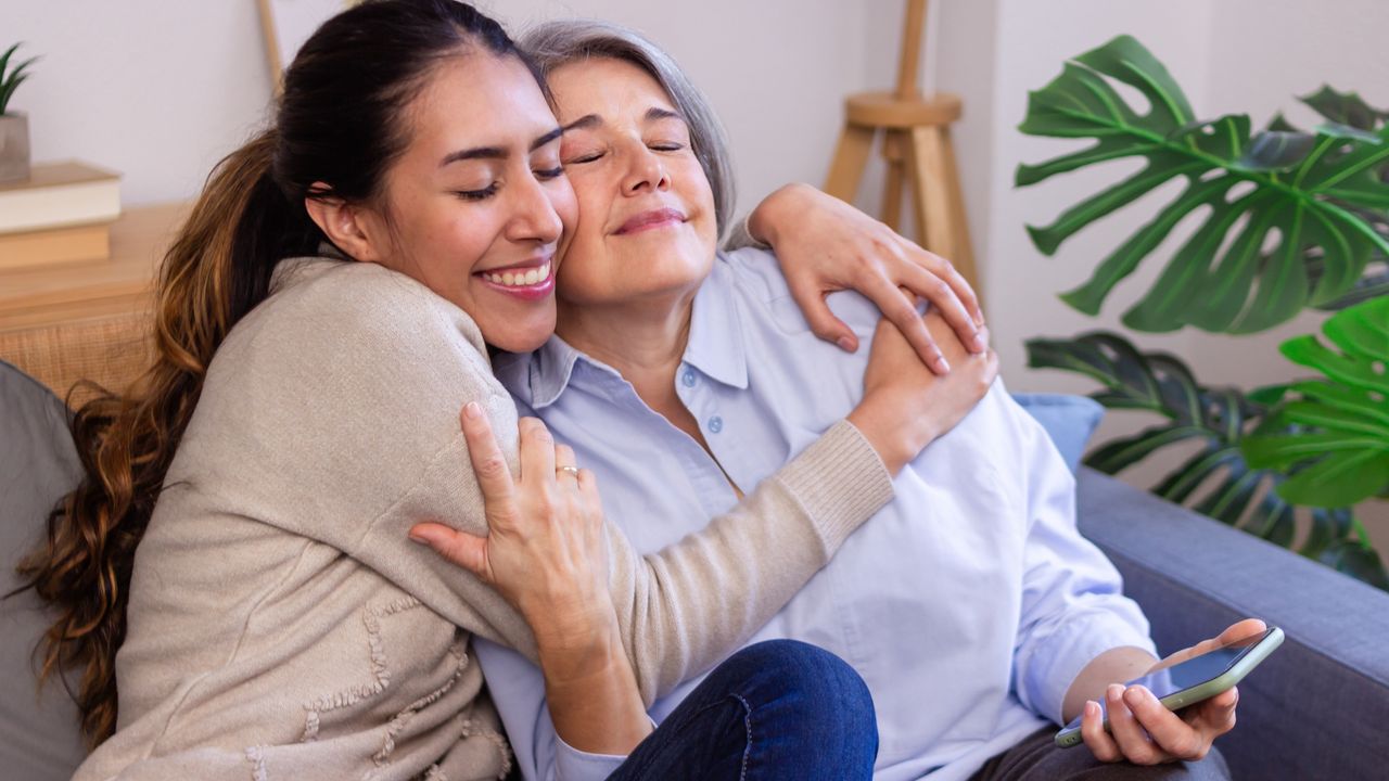 An adult daughter hugs her smiling mom on the sofa.