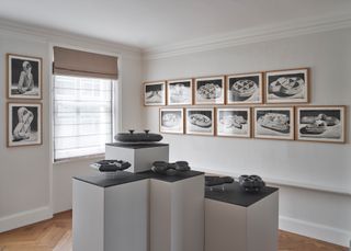 Interior view of a room with white walls, a window, wood flooring, black and white framed artworks on the walls and concrete pieces on black and white plinths by Alexandre Arrechea at SoShiro