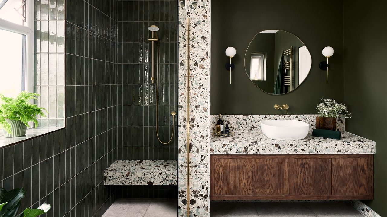 a green bathroom with a vanity area with a single white basin, terrazzo wall and countertop, wooden floating vanity unit, globe wall lights and round wall mirror beside a shower area with green metro tiles and gold fixtures