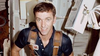 a smiling man in a blue shirt sits inside the space shuttle, surrounded by equipment