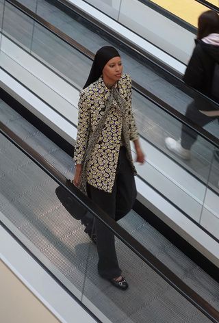 A winter travel outfit is shown in a photo of a woman walking in an airport terminal wearing a black hijab with a black and yellow floral print coat, skinny printed neck scarf, black sweater, black relaxed jeans, black ballet flats, and black Gucci bag