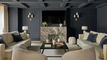 Black living room with cream sofas and armchairs, black coffee table, beige and brown marble fireplace, wall-mounted TV, three-pronged wall lights and cream pouffe