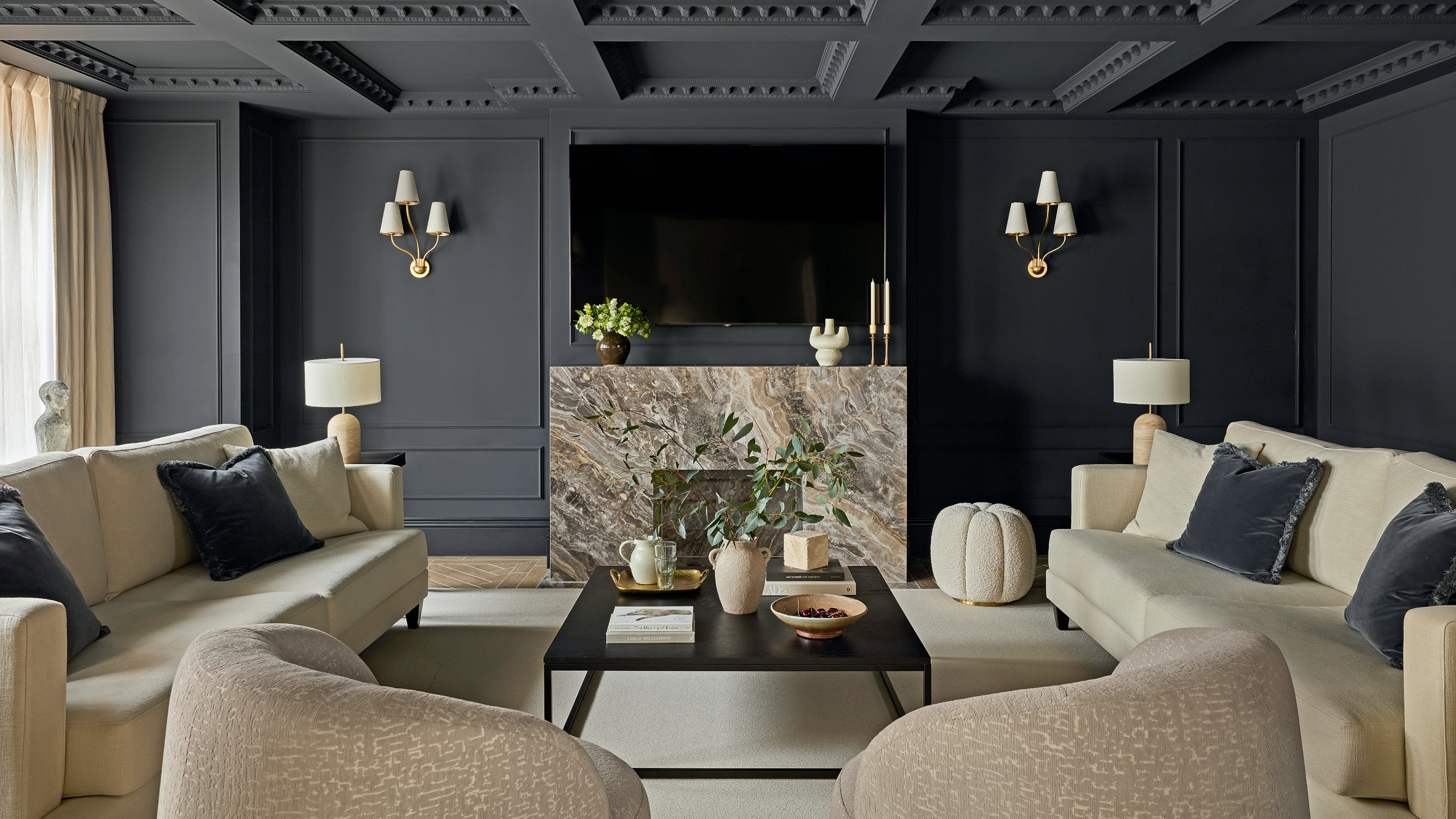 Black living room with cream sofas and armchairs, black coffee table, beige and brown marble fireplace, wall-mounted TV, three-pronged wall lights and cream pouffe