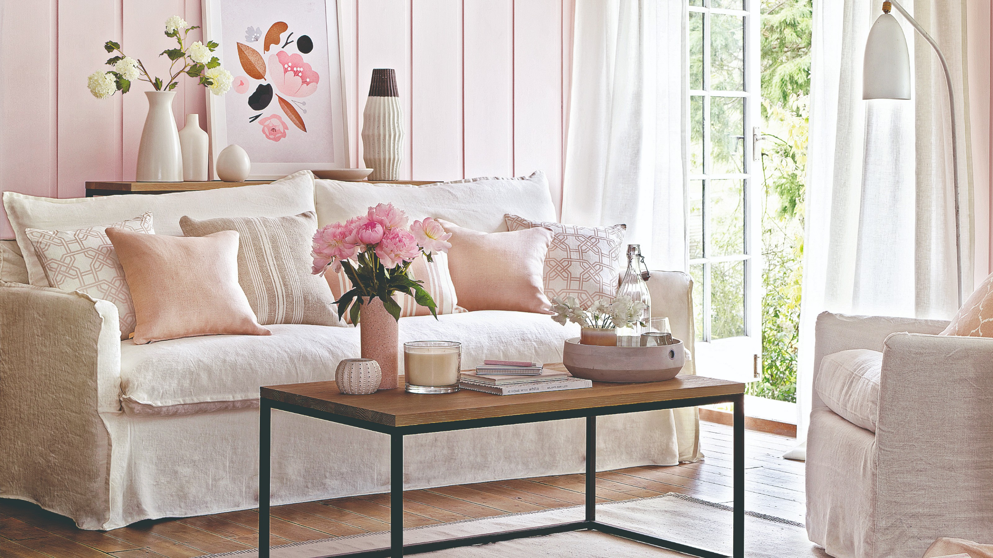 A pink living room with a white sofa and an open glass door to the garden with a vase of pink flowers and a large scented candle on the coffee table