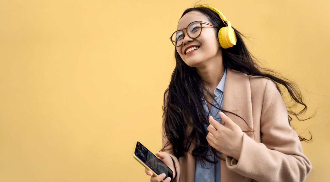 Young asian woman in coat with yellow headphones and mobile phone listening to music via bluetooth and dancing near the wall on the street. Mental and physical health. Headspace vs. Calm, meditation apps