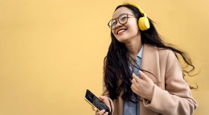 Young asian woman in coat with yellow headphones and mobile phone listening to music via bluetooth and dancing near the wall on the street. Mental and physical health. Headspace vs. Calm, meditation apps