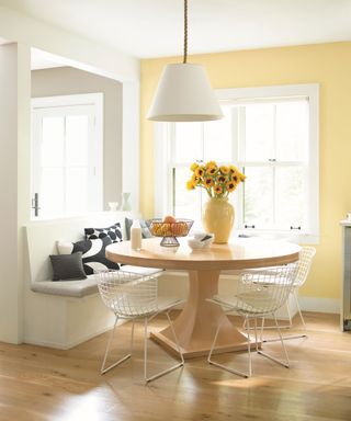 Bright dining room with white painted walls and light yellow painted feature wall, light wooden rounded dining table, white wired dining chairs, built in dining bench with seat cushions, light wooden flooring, white pendant hanging over table