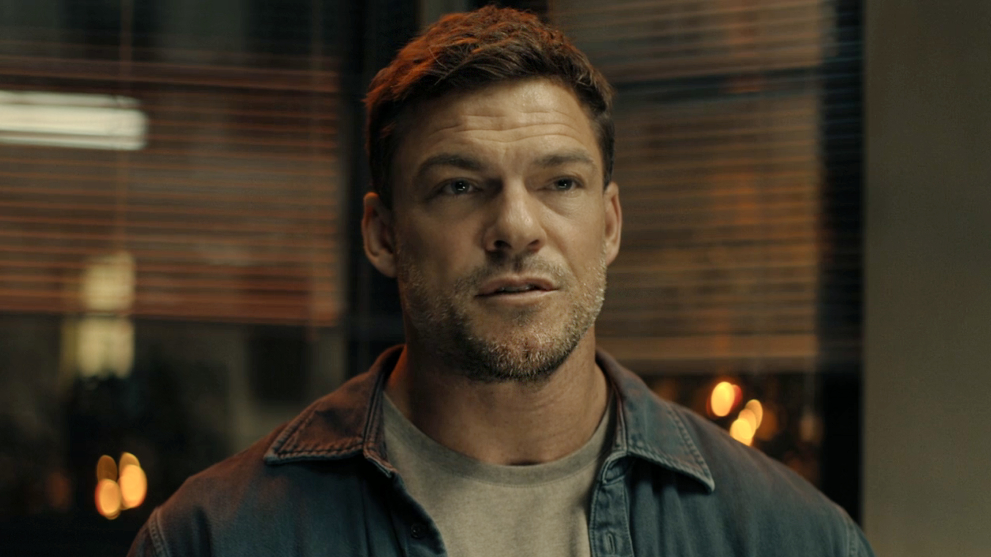 Alan Ritchson, wearing grey shirt and open button up denim over shirt, sits in a police interrogation room in REACHER Season 4 Episode 1.