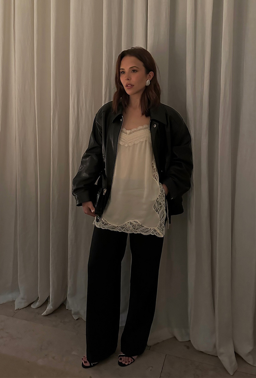 A woman standing inside against a white curtain backdrop wearing an oversize leather jacket over a white sheer lace camisole, dark wash jeans, and black strappy sandals.