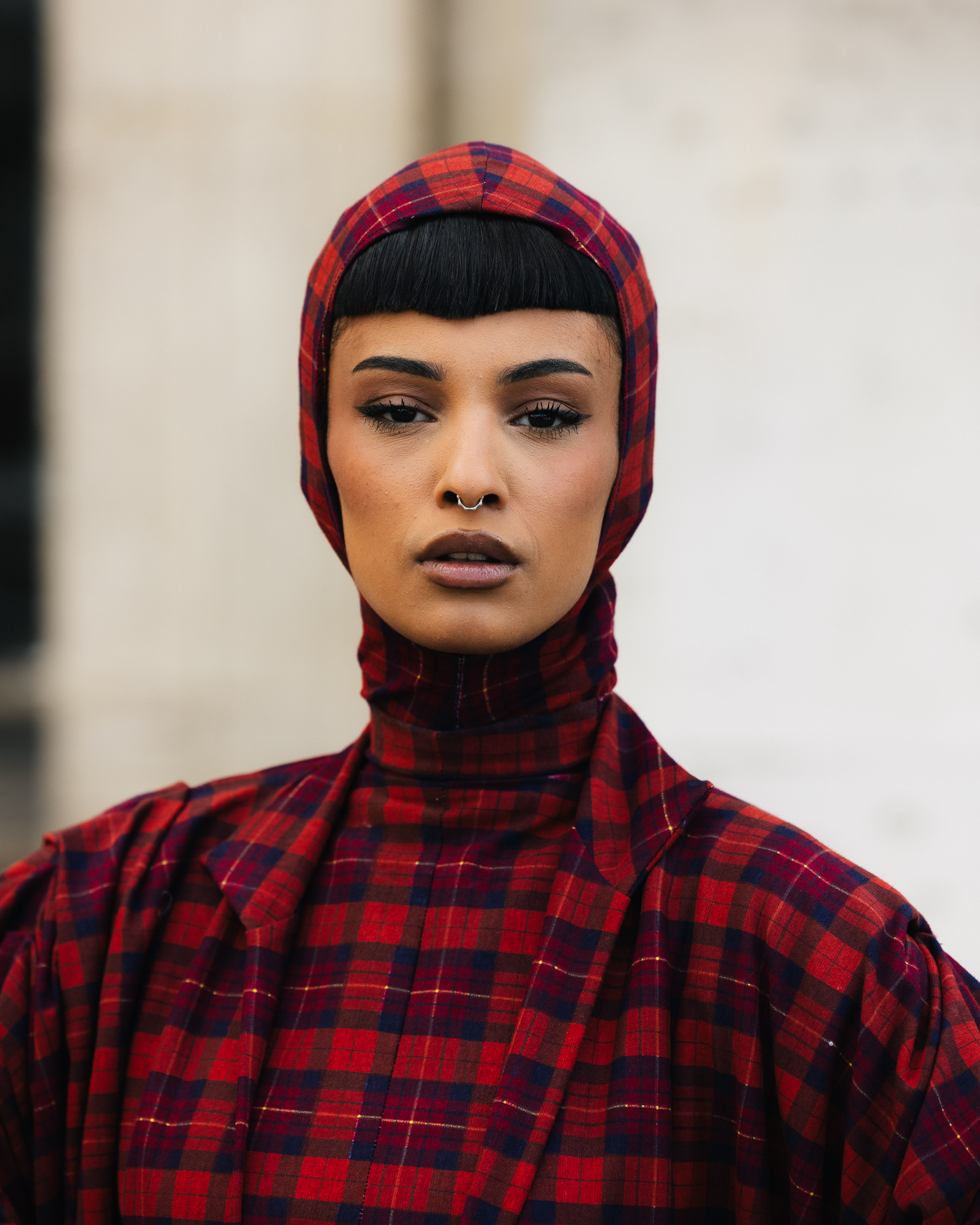 a woman with blunt bangs and a septum ring wearing a red plaid hooded jacket