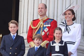 Prince George, Prince Louis, Princess Charlotte, Prince William and Princess Kate standing on the Buckingham Palace balcony in dress clothes and suits smiling