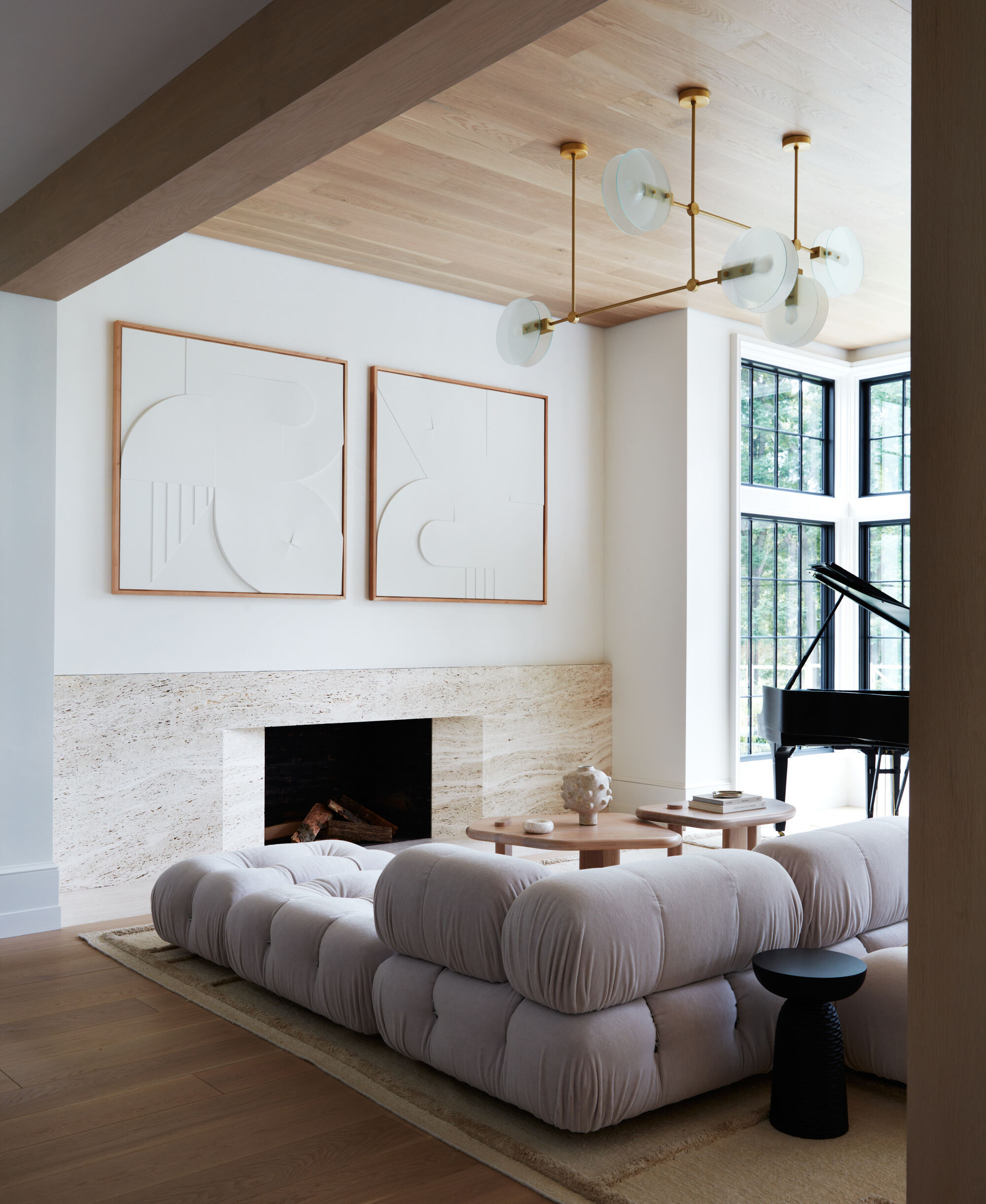 beige living room with travertine slab fireplace, two sculptural white artworks hung above, a timber clad ceiling, pendant light, piano in the corner surrounded by steel-framed windows, and a B&B camaleonda sofa