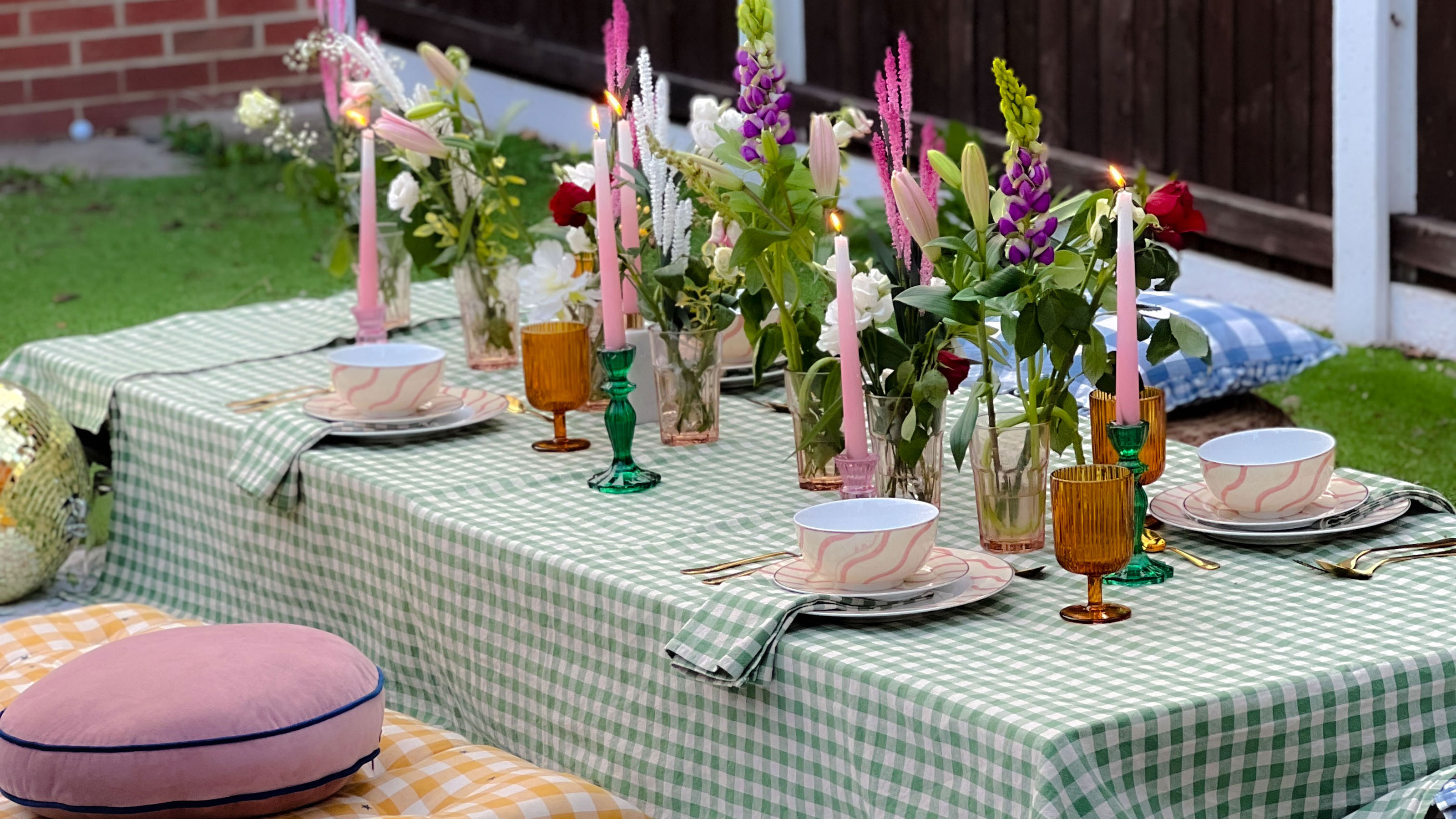 Outdoor table with green and white gingham tablecloth, flowers in tumblers and flatware with pink and white squiggle pattern alongside yellow and white gingham floor cushion and round pink pillow