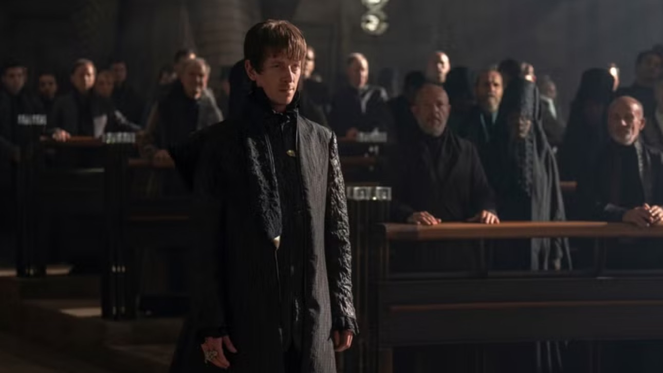 a man in a dark tunic stands in a courtroom full of people dressed in black