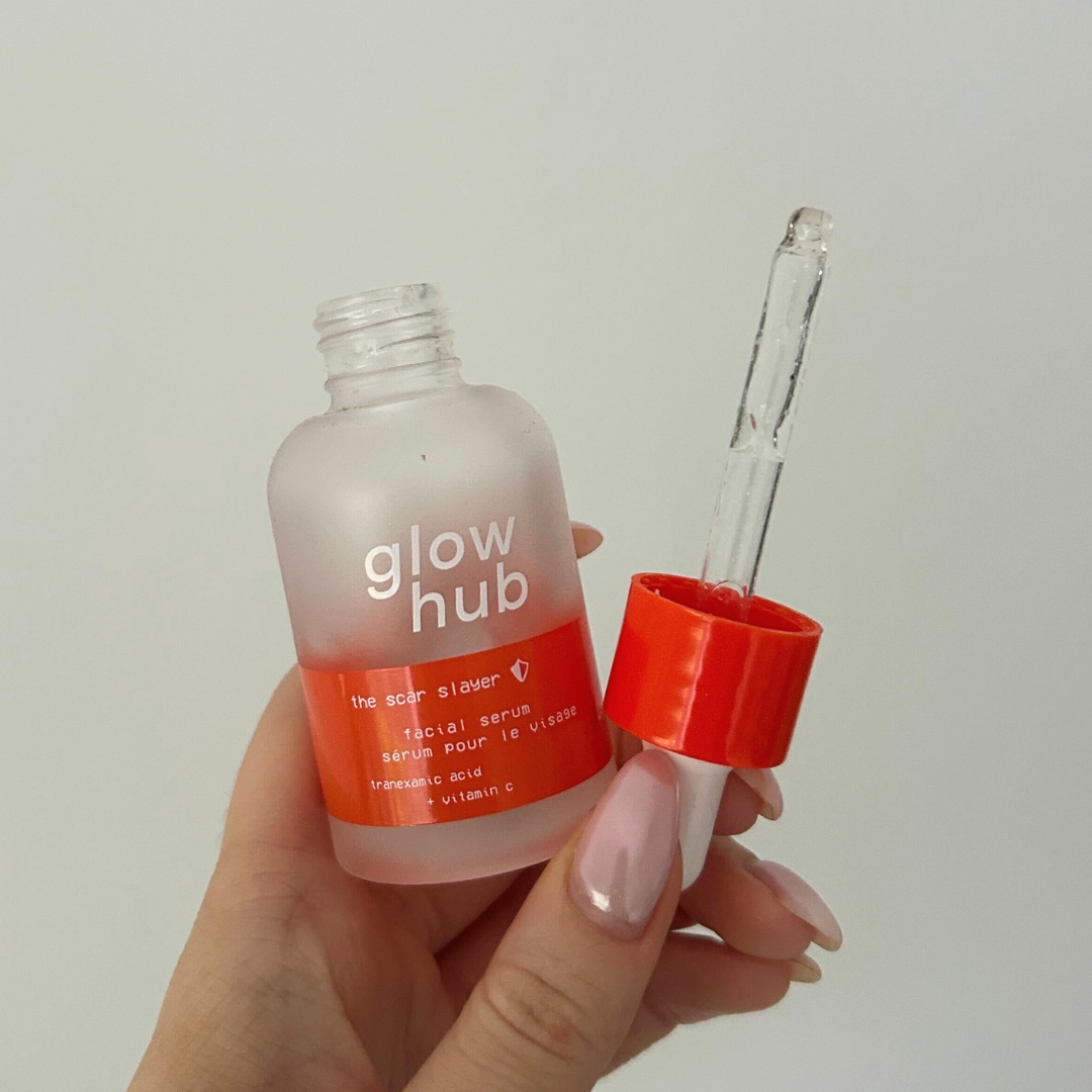 Hand holding an empty bottle of the Glow Hub The Scar Slayer Facial Serum against a white wall