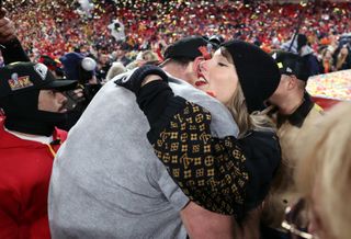 Taylor Swift wearing a black Louis Vuitton hoodie and matching winter hat hugging Travis Kelce, wearing a gray t-shirt, as confetti falls in the air on the field after the AFC 2025 Championship.