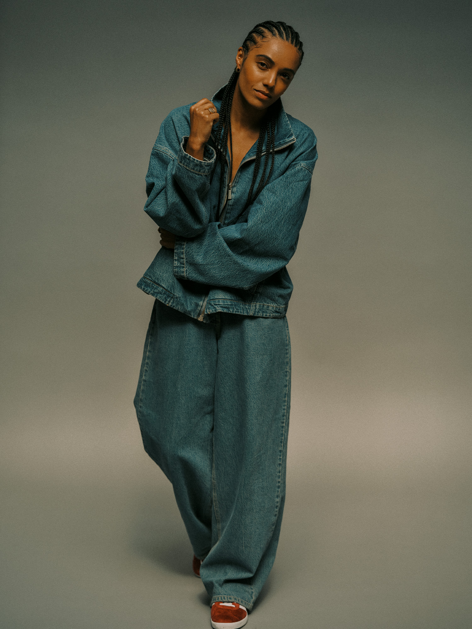 Actor Maisie Richardson-Sellers poses on a gray seamless backdrop wearing an oversize zip-up denim jacket and baggy jeans with red Adidas sneakers.
