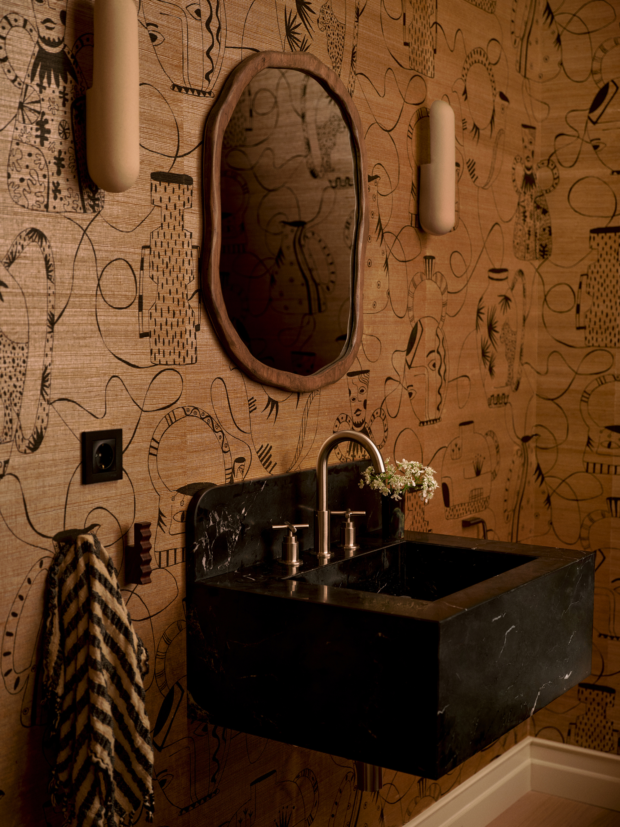 neutral powder room with black marble sink, brass tapware, patterned timber walls, round wooden mirror, wall sconces, striped hand towel on hook