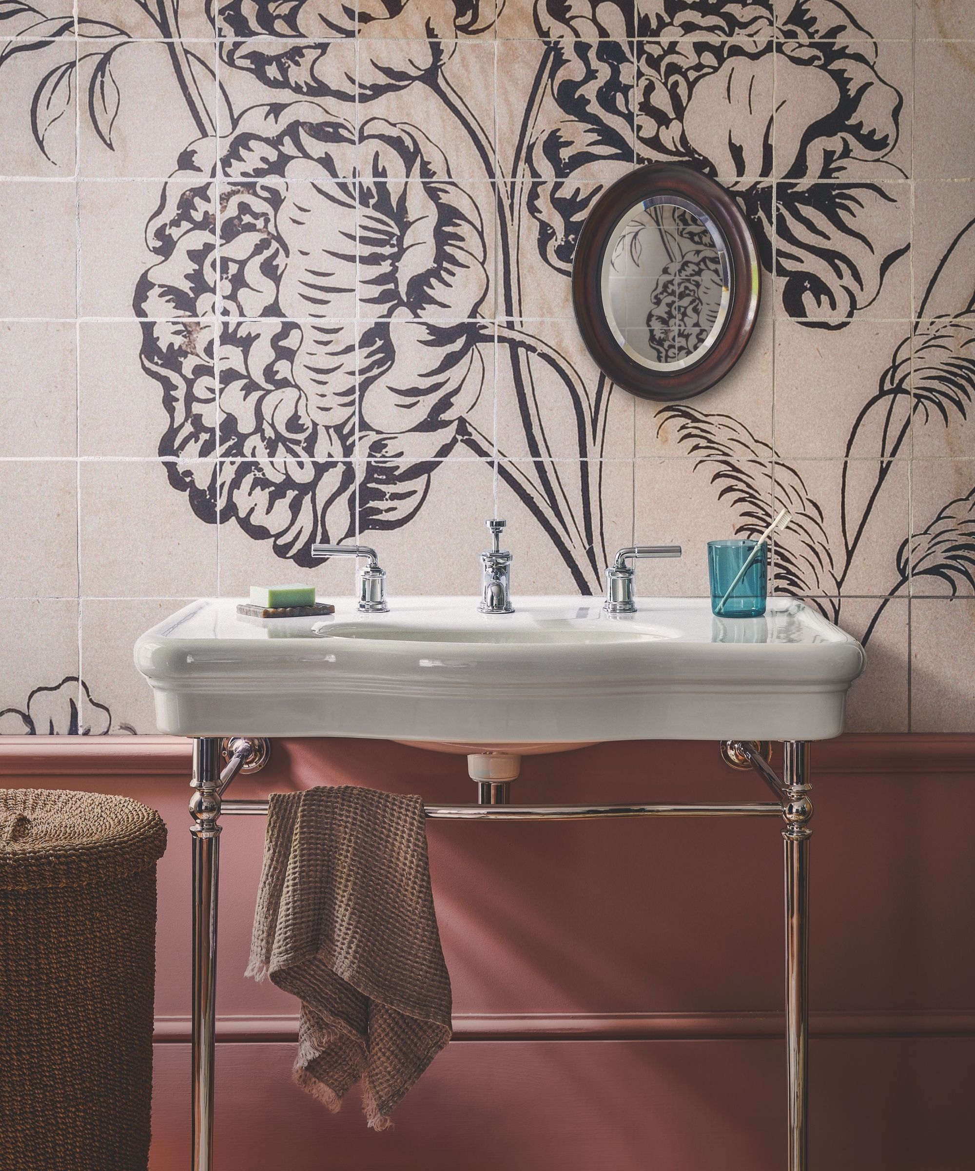 Terracotta bathroom with floral mural tiles, oval mirror with dark brown frame, white sink with silver legs and faucets, brown towel, and a brown woven laundry hamper.