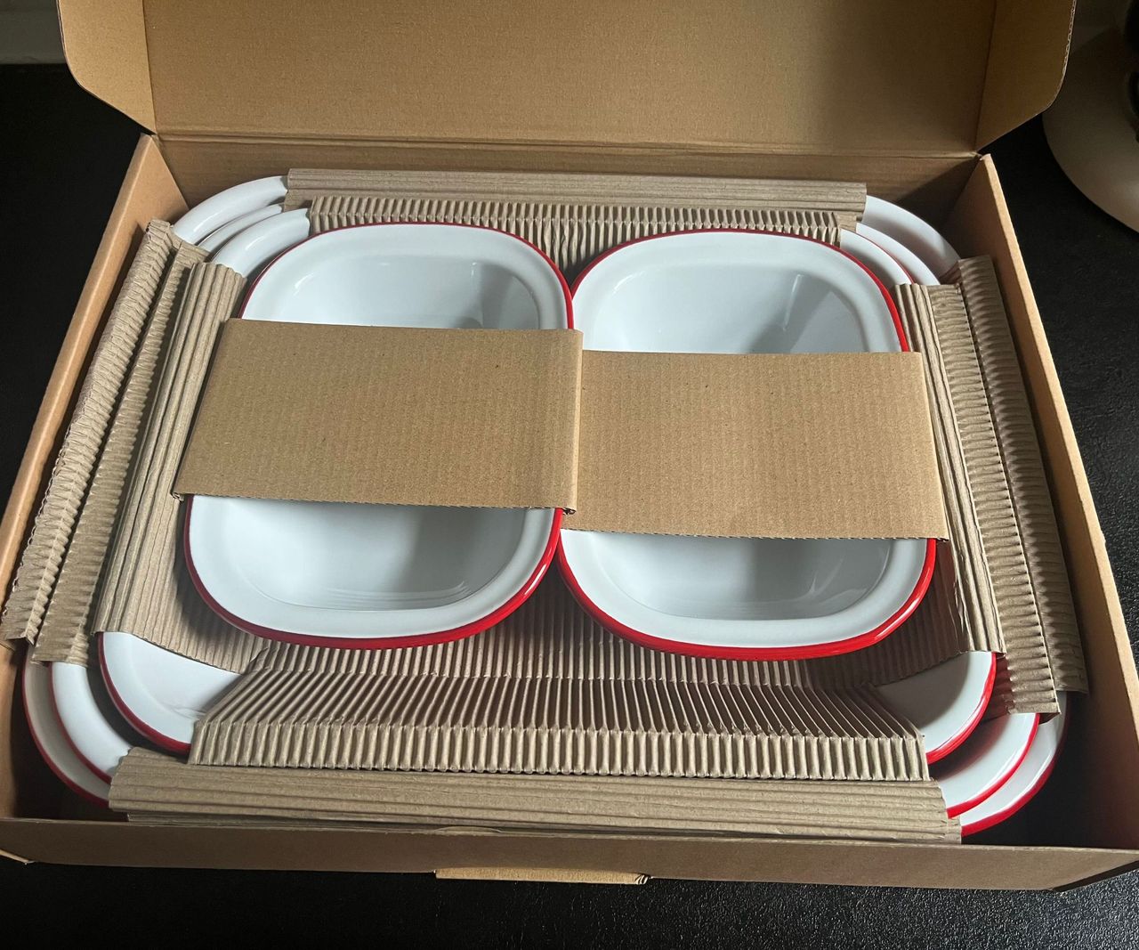 A set of white enamel dishes with red rims, inside a cardboard box.