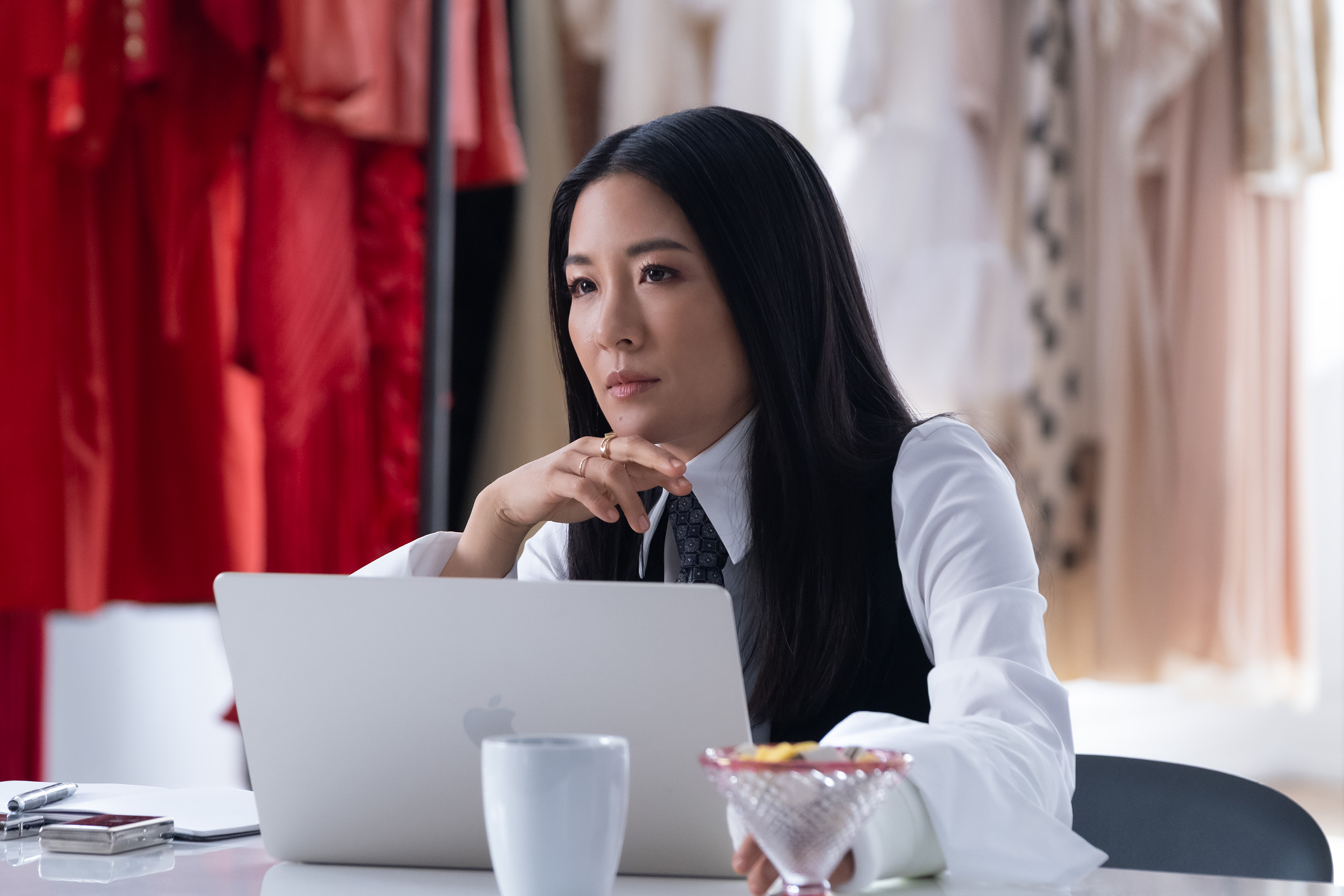 constance wu as vanessa wearing a vest and tie sitting at a laptop in front of clothes in not suitable for work
